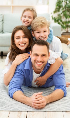 Positive Family Of Four Lying Posing On Floor Indoors. Weekend Together At Home. Vertical